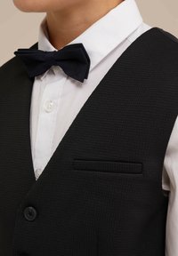 Svart texturerad väst med en vit skjorta under, som har en svart fluga och en liten bröstficka.