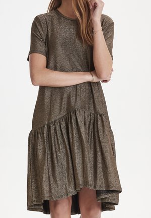 Woman wearing a shimmery bronze short-sleeve dress with an asymmetrical gathered hem, standing with arms crossed and hand near her face.