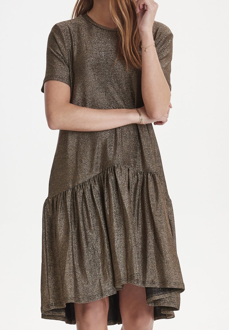 Femme portant une robe courte à manches courtes bronze scintillante avec un ourlet asymétrique froncé, debout les bras croisés et la main près du visage.