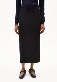 Zwarte, strak aansluitende, enkellange rok van een gladde stof. Gecombineerd met zwarte schoenen met een bandje en ronde hardware-accenten.