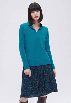 Maglione a maglia turchese con colletto a polo, abbinato a una gonna a stampa floreale scura. Tessuto morbido, vestibilità rilassata e bordo a coste discreto.
