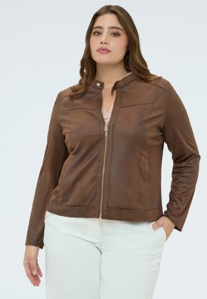 Femme portant une veste marron à fermeture éclair avec un col montant et un pantalon blanc, debout avec une main dans la poche, sur un fond uni.