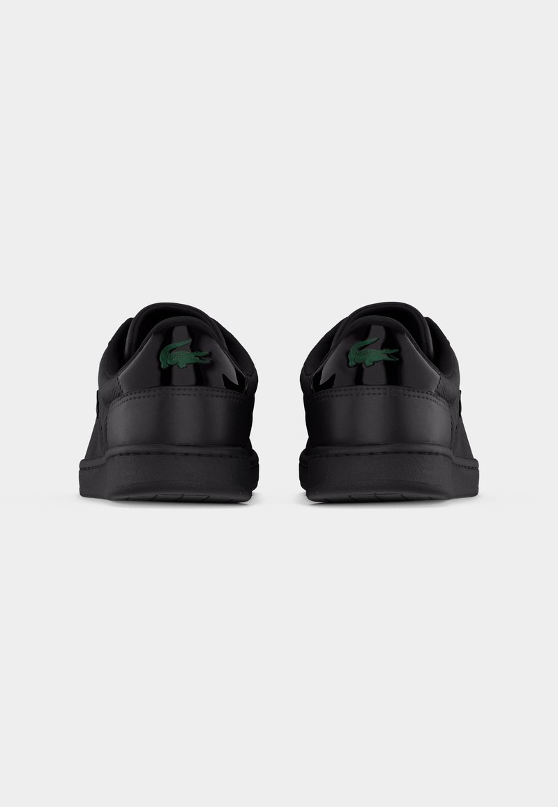 Coppia di sneaker nere basse viste da dietro, con un logo di coccodrillo verde sulla linguetta del tallone, su uno sfondo chiaro e uniforme.