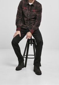 Chemise à manches longues à carreaux bruns avec des accents rouges sur les poches, associée à un pantalon noir et des bottes noires, assise sur un tabouret.