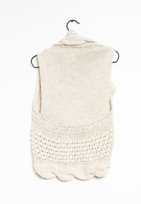 Gilet senza maniche lavorato a maglia appeso a un gancio nero. Di colore crema con motivi testurizzati nella parte inferiore, caratterizzato da un bordo ondulato.