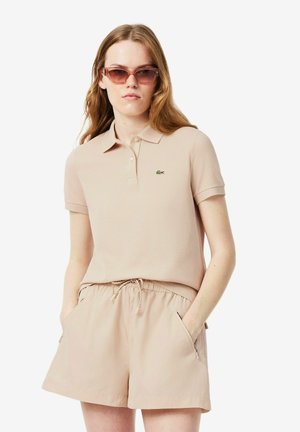 Vrouw met lang haar die een beige poloshirt draagt, bijpassende shorts met trekkoord, handen in de zakken en een bruine zonnebril op een witte achtergrond.