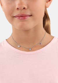 Silberne Kettenhalskette mit drei Anhängern: einem Schmetterling, einer Blume und einem Herz, die jeweils mit bunten Akzenten verziert sind. Tragen über einem pinken Shirt.