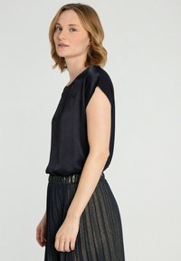 Navyblauwe satijnen blouse met korte, opgerolde mouwen en een losse pasvorm, gecombineerd met een geplooide, glinsterende donkere rok. Eenvoudig en elegant ontwerp.