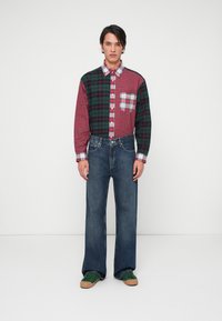 Camicia a quadri rossa e verde con tasca, abbinata a jeans larghi in denim scuro e sneakers verdi. Design semplice e casual con motivi a contrasto.