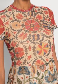 Robe à motifs floraux avec des designs complexes en rouge, jaune et bleu sur un fond beige, avec un col rouge uni et des manches courtes.