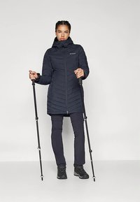 Sieviete, tērpusies melnā kapucēta ziemas mētelī, tumšās biksēs, pārgājienu zābakos, turot melnus pārgājienu nūjas, stāv uz vienkārša gaiša fona.