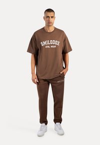 Camiseta de algodón marrón de gran tamaño con impresión blanca "SMILODOX ATHL. WEAR", combinada con joggers marrones a juego y zapatillas blancas.