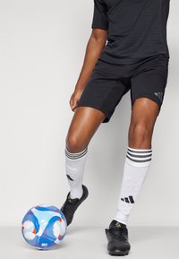 Zwart sportshirt en -short, zwarte voetbalschoenen, witte sokken met zwarte strepen. Een blauw en rood gepatrooneerde voetbal ligt op de grond.