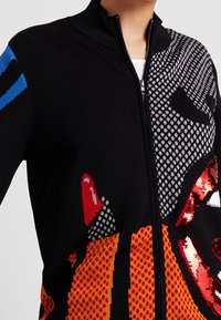 Person som bär en svart zip-up tröja med färgglada geometriska och prickiga mönster i rött, orange, blått och vitt, inklusive glänsande röda paljetter.