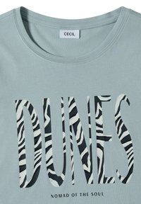 Chemise bleu clair avec un grand texte imprimé "DUNES" en rayures zèbre noires et blanches et un texte plus petit en dessous disant "NOMAD OF THE SOUL."