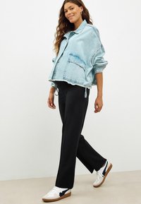 Jachetă oversized din denim albastru deschis cu margine brută, pantaloni negri și adidași albi cu accente închise și talpă texturată.