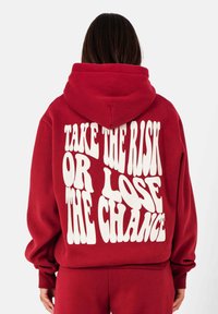 Rode hoodie met grote witte tekst op de achterkant die zegt "NEEM DE RISK OF VERLOST DE KANS." Zachte stof en oversized pasvorm.