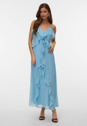 Kobieta w jasnoniebieskiej, bezrękawnikowej sukience maxi z kaskadowymi falbanami z przodu, w połączeniu z czarnymi sandałami na obcasie z odkrytymi palcami.