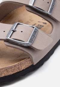 Εγγύς λήψη μπεζ σανδαλιού Birkenstock με δύο ρυθμιζόμενα λουράκια και μεταλλικές αγκράφες πάνω σε φελλό πάτο και μαύρη σόλα.