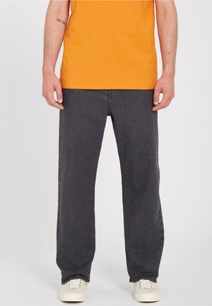 Homme portant un jean droit gris foncé, des baskets blanches et une chemise orange, debout devant un fond blanc uni.