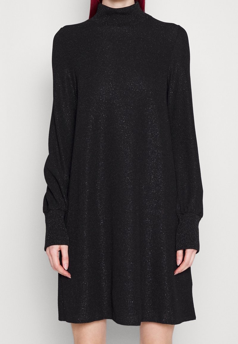 Femme portant une robe en maille noire scintillante à manches longues avec un col montant sur un fond uni gris clair.