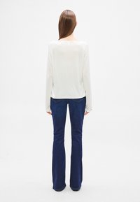 Hosszú ujjú, fehér felső széles nyakkivágással és sima textúrával, sötétkék, bővülő szárú farmerral párosítva, amely ötpockets kialakítású és testhez simuló.