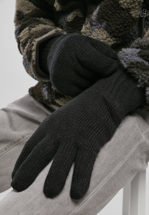 Gants en tricot noirs avec texture côtelée, présentant un design ajusté. Portés par une personne assise, partiellement couverte par une veste en polaire à motifs.