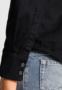 Svart denimskjorta med knappdetalj vid ärmslut, synlig textur och en avslappnad passform, kombinerad med ljusblå denimjeans.