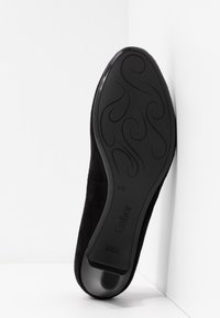 Semelle noire d’une chaussure femme avec motifs décoratifs et nom de marque Gabor, positionnée verticalement dans un coin blanc.