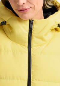 Geel pufferjack met een hoge kraag en een zwarte rits. Kenmerken zijn onder andere een gewatteerde textuur en verstelbare hardware op de capuchon.