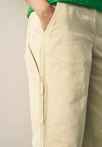 Beige cargo-housut, joissa on sivutasku ja sileä pinta. Yhdistetty vihreän neulepuseron kanssa, näyttäen läheltä vaatteen yksityiskohtia.