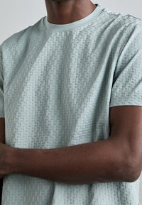 Krátký rukáv, texturované tričko v světle mátové barvě, s jemným tkaným vzorem, kulatým výstřihem a volným střihem.