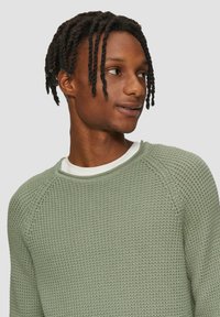 Hellgrüner Strickpullover mit strukturierter Musterung, Rundhalsausschnitt und gerippten Manschetten, übereinander getragen mit einem weißen Hemd.