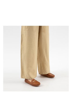 Beige wijde broek gecombineerd met bruine geperforeerde loafers met een gouden gesp op een witte achtergrond.