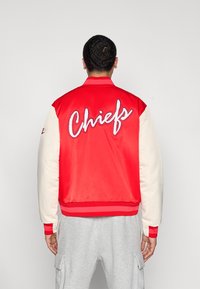 Κόκκινο και κρεμ varsity σακάκι με κεντημένο το "Chiefs" στην πλάτη. Διαθέτει λεία σατέν επιφάνεια και ριμπ μανσέτες και τελείωμα.