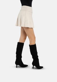 Botas negras de ante hasta la rodilla con diseño holgado y punta afilada, combinadas con una minifalda plisada beige que muestra una silueta ajustada.