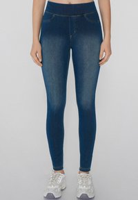 Mujer vestida con jeans skinny azul oscuro y zapatillas blancas, de pie frente a un fondo claro y liso.