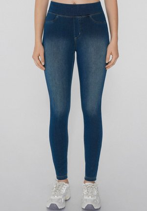 Mujer vestida con jeans skinny azul oscuro y zapatillas blancas, de pie frente a un fondo claro y liso.