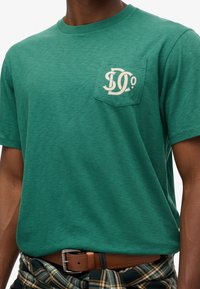 Camiseta de algodón verde con un bolsillo en el pecho que presenta un diseño de logo en crema; mangas cortas y cuello redondo; textura suave.