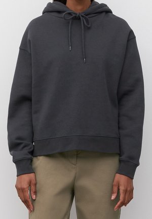 Osoba ubrana w ciemnoszarą bluzę z kapturem i ściągaczami oraz beżowe spodnie, stojąca na jednolitym jasnym tle.