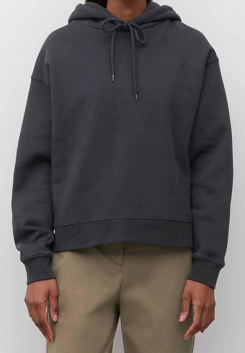 Personne portant un sweat à capuche gris foncé avec des cordons de serrage et un pantalon beige, debout devant un fond clair uni.