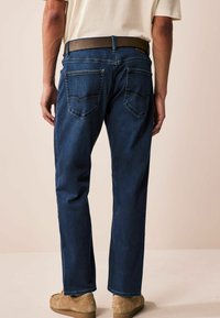Jeans din denim de culoare albastră închisă, cu un design drept, cinci buzunare și cusături contrastante, asortate cu o curea maro și pantofi de culore deschisă.