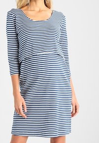 Kvinna som bär en blå och vit horisontellt randig klänning med tre fjärdedels ärmar, stående med händerna avslappnade längs sidorna.