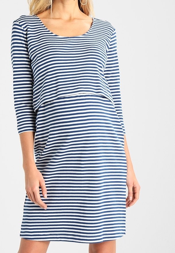 Kvinna som bär en blå och vit horisontellt randig klänning med tre fjärdedels ärmar, stående med händerna avslappnade längs sidorna.