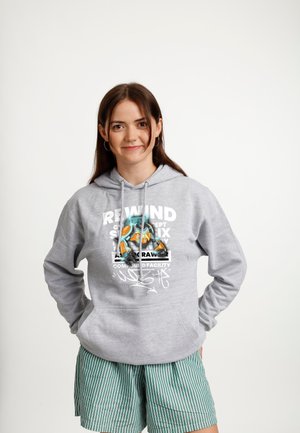 Młoda kobieta uśmiechająca się, ubrana w szarą bluzę z kolorowym nadrukiem oraz zielone szorty w paski, na jednolitym białym tle.