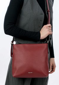 Borsa a spalla rossa in pelle con finitura tessurizzata, dotata di chiusura con zip e tracolla regolabile, tenuta in mano su uno sfondo grigio.