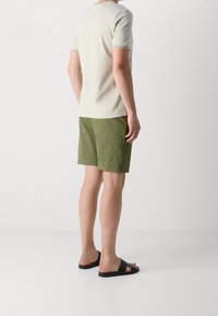 T-shirt leggero beige a maniche corte, abbinato a pantaloni corti verde oliva e sandali neri. La maglietta ha un colletto rotondo e cuciture a contrasto.