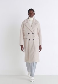 Cappotto lungo di colore beige chiaro, doppiopetto, realizzato in un tessuto testurizzato, con ampi revers, due tasche frontali e bottoni scuri. Indossato sopra un dolcevita bianco.