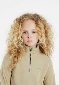 Béžový fleece pulóver s vysokým golierom a polovičným zipsom. Obsahuje jemné logo v zodpovedajúcej farbe. Mäkká textúra a kučeravé vlasy rámujúce tvár.