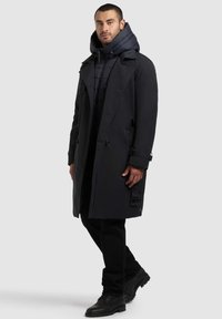 Tume trench-coat eemaldatava kapuutsiga, kihitatud musta polsterdatud jope üle. Omab ees nuppe, küljetaskuid ja sujuvat tekstuuri.